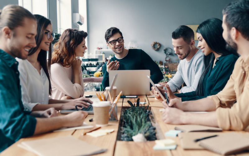 A diverse group of individuals gathered around a table, engrossed in their work on a laptop.